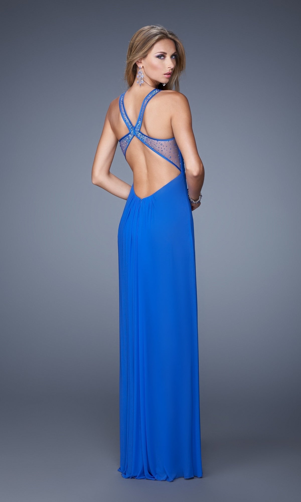 A woman wears the floor-length La Femme Dress 20903 in bright blue, featuring an open back with crisscross embellished straps, standing against a plain gray background.